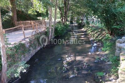 Petit ruisseau bordé de végétation avec des tentes blanches installées pour un événement en arrière-plan.