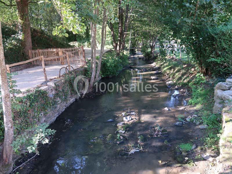 Ruisseau bordé d'arbres et de végétation, avec un chemin en gravier et une barrière en bois sur la gauche.