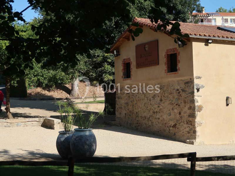 Façade d'un bâtiment en pierre et crépi avec des pots décoratifs, entouré de végétation et d'une allée en gravier.