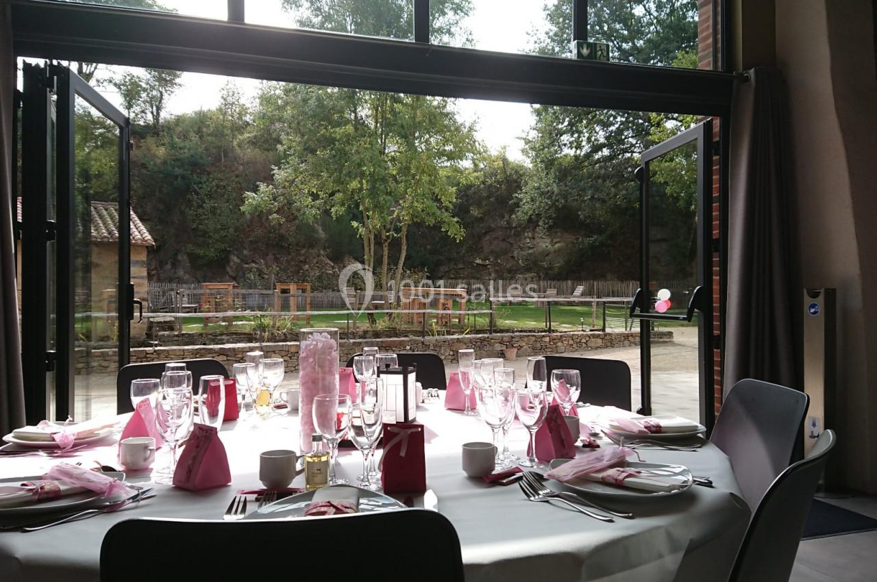 Table dressée avec nappes blanches et serviettes roses, disposée près de baies vitrées ouvertes sur un jardin verdoyant.
