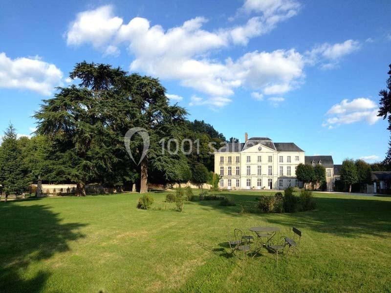 Manoir entouré d'un grand jardin verdoyant avec des arbres imposants sous un ciel bleu parsemé de nuages.