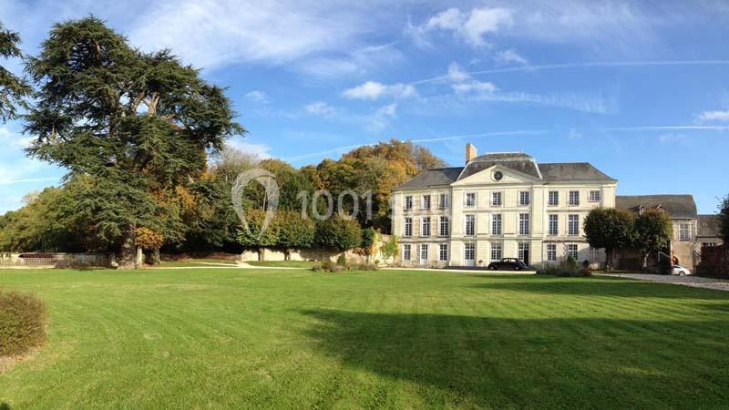 Manoir blanc entouré d'un grand jardin verdoyant, avec des arbres et un ciel bleu en arrière-plan.