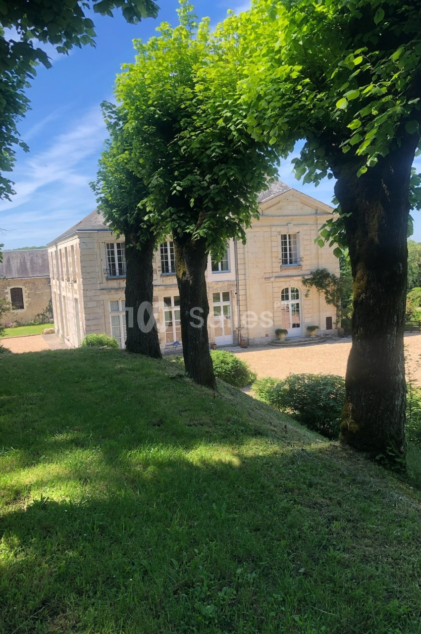 Façade d'une maison en pierre entourée d'arbres et de pelouse, sous un ciel bleu dégagé.