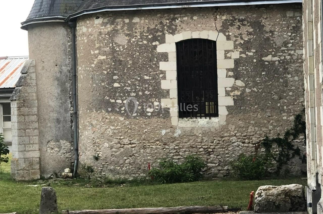 Mur en pierre d'un bâtiment ancien avec une fenêtre à barreaux, entouré d'herbe et de végétation.
