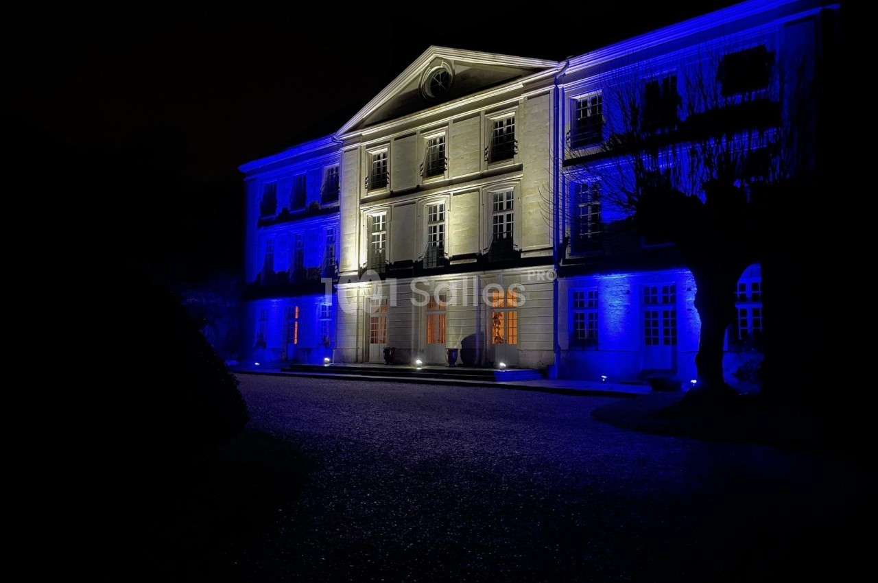 Façade d'un bâtiment éclairé en bleu et blanc la nuit, avec des arbres et une allée au premier plan.