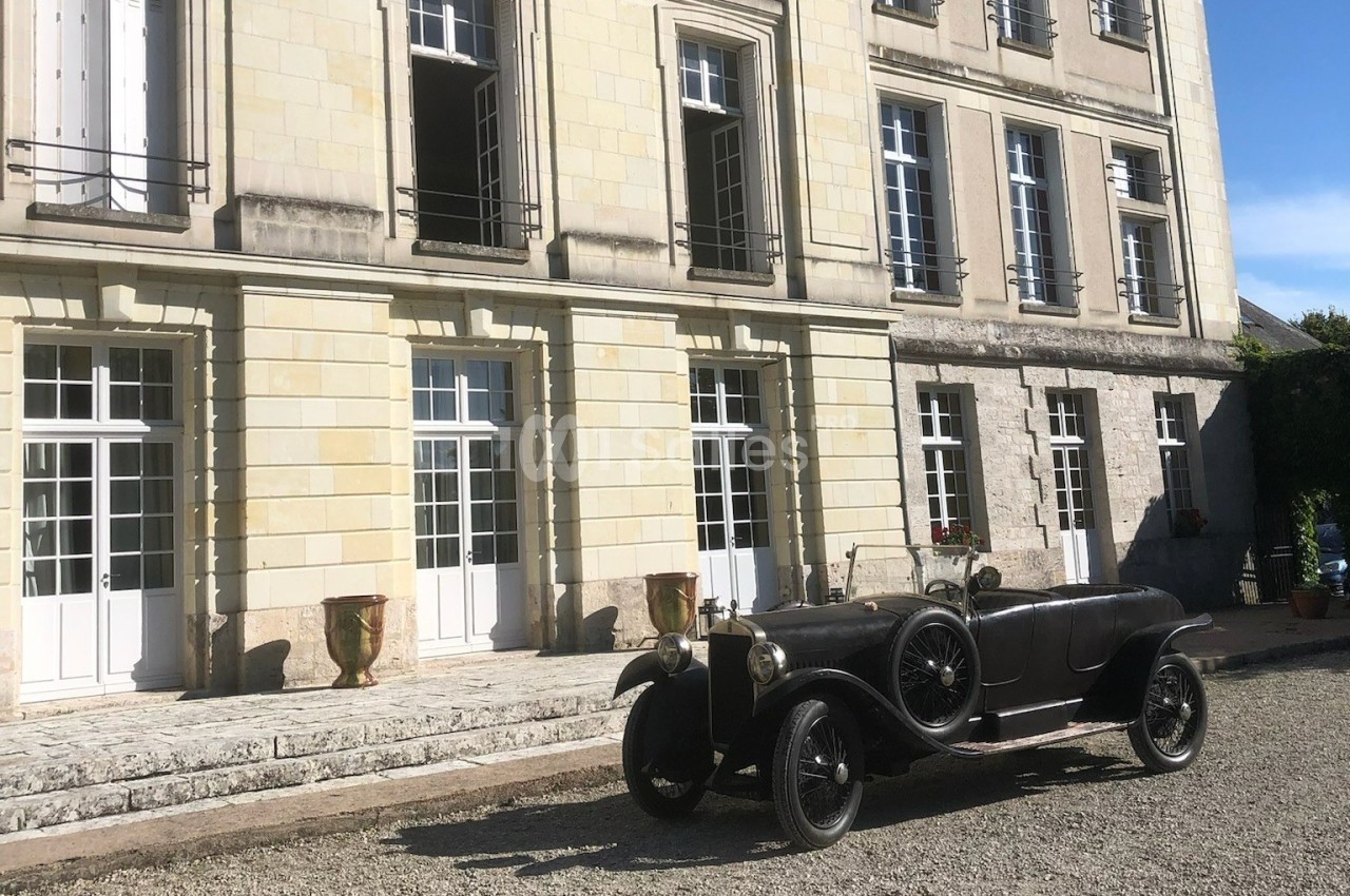 Voiture ancienne noire stationnée devant un bâtiment en pierre avec de grandes fenêtres et des jardinières.