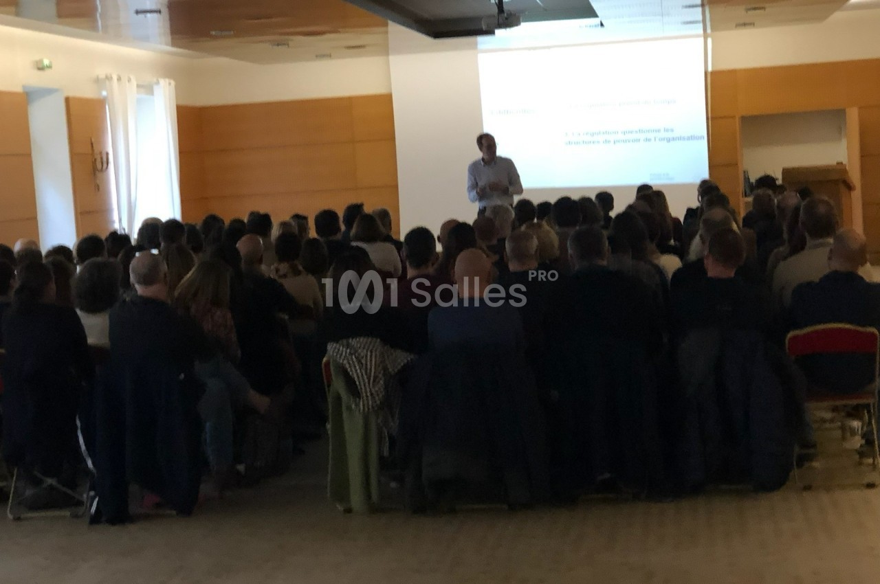 Un conférencier présente devant un public assis dans une salle équipée d'un écran de projection.