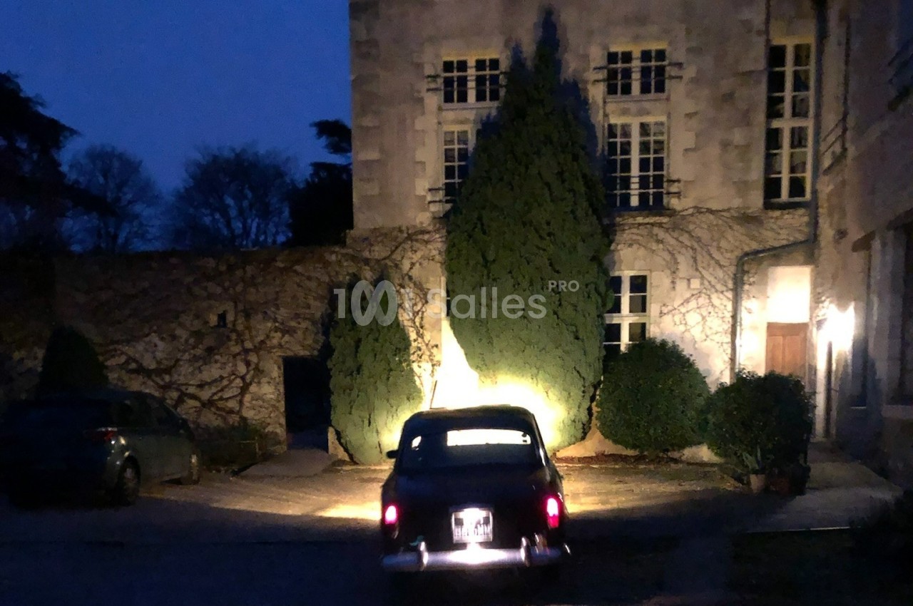 Voiture ancienne garée de nuit devant un bâtiment en pierre éclairé, entouré de végétation.