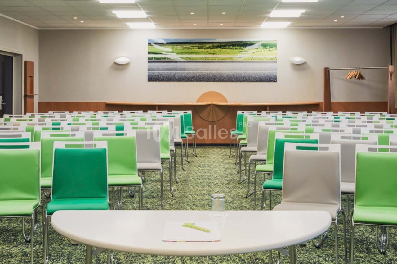 Salle de conférence lumineuse avec des rangées de chaises vertes et blanches, table blanche au premier plan.
