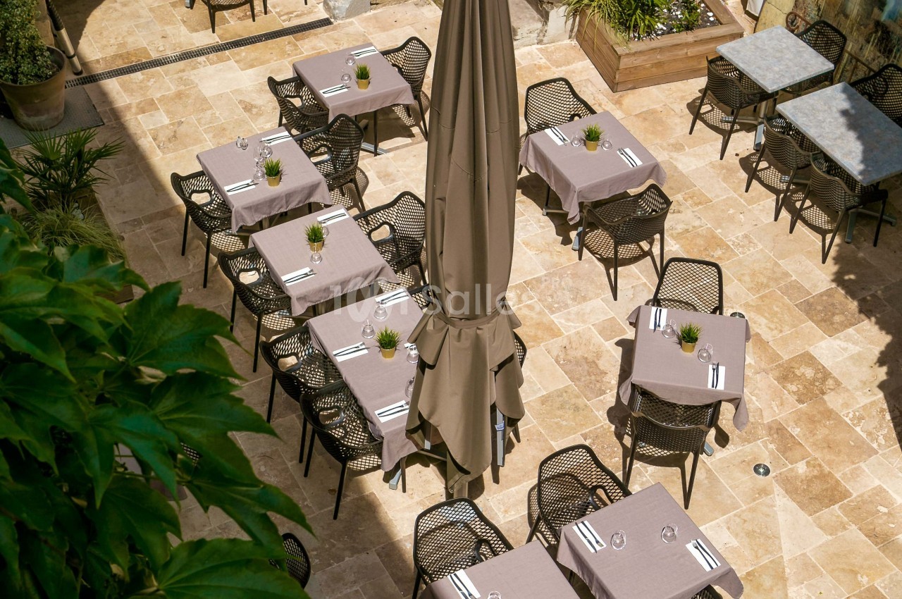 Terrasse d'un restaurant avec des tables dressées, nappes grises, chaises noires et un grand parasol central.
