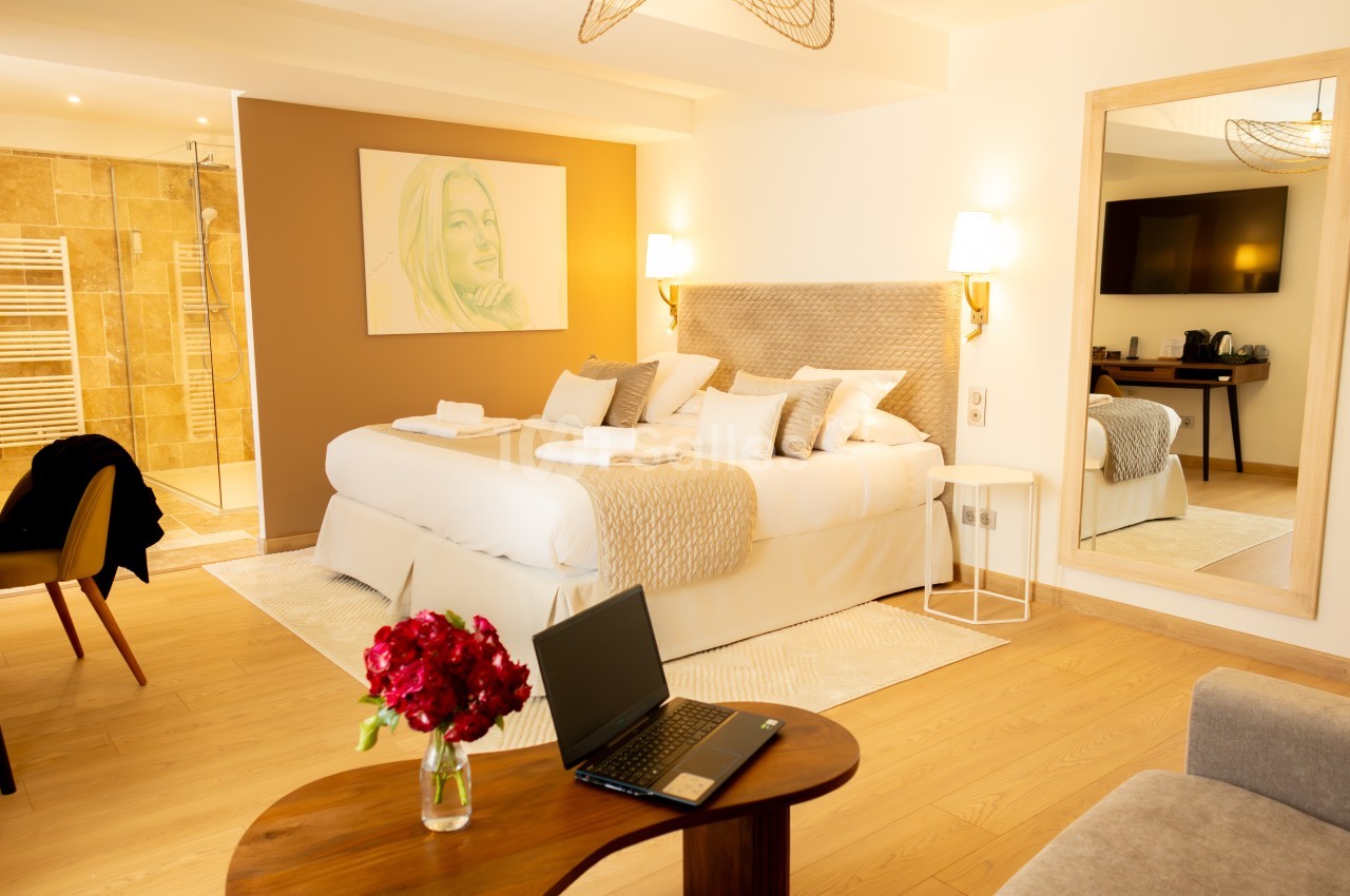 Chambre lumineuse avec lit double, salle de bain attenante, bureau avec ordinateur portable et vase de fleurs rouges.
