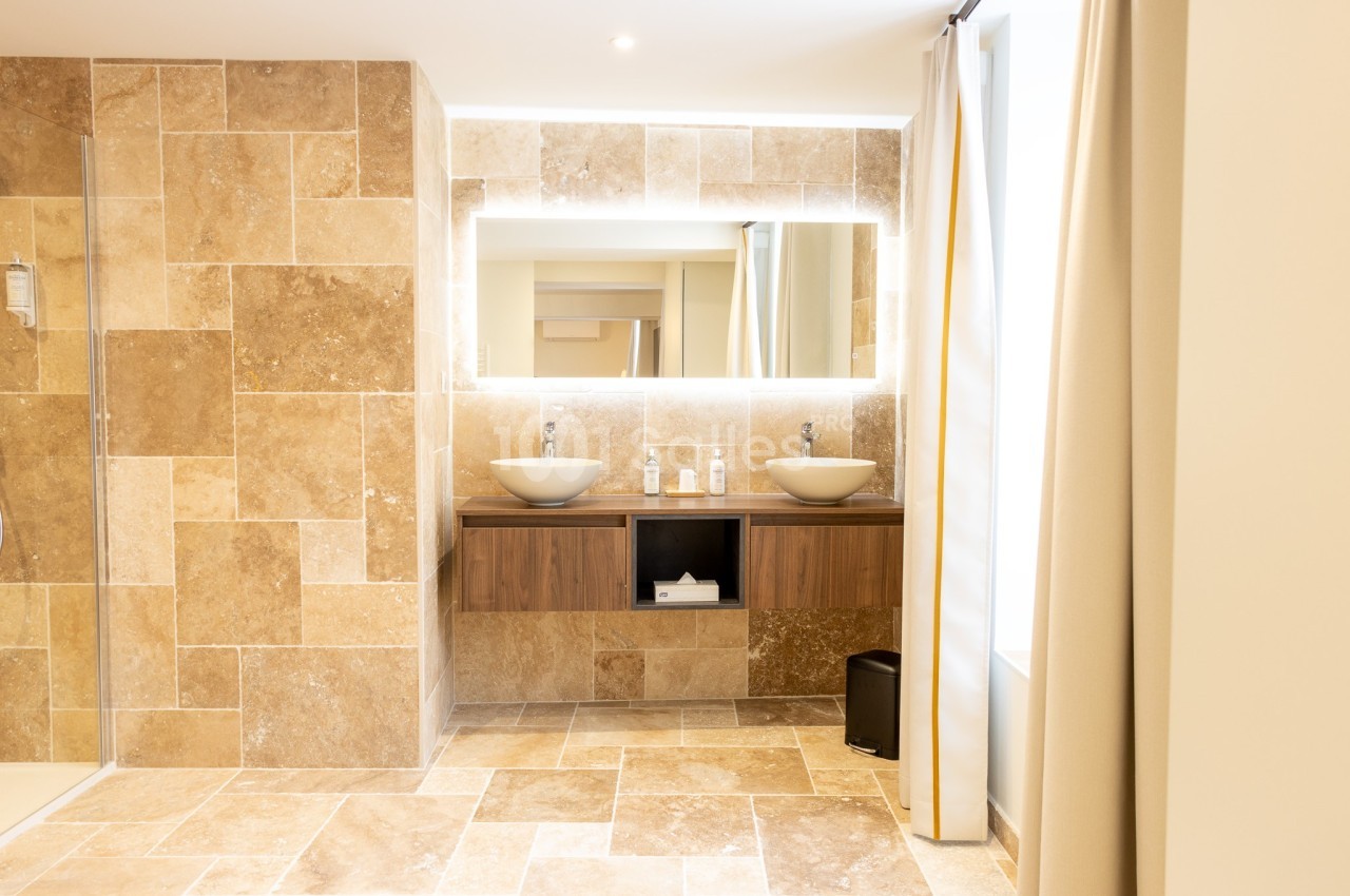 Salle de bain moderne avec deux vasques, un grand miroir éclairé et des murs en carrelage beige.