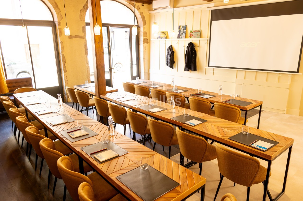 Salle de réunion lumineuse avec des tables en bois, des chaises alignées et un écran de projection à l’arrière.