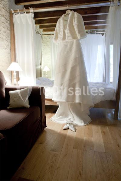 Robe de mariée blanche suspendue dans une chambre lumineuse avec murs en pierre et poutres apparentes.