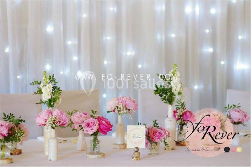 Table décorée avec des fleurs roses et blanches dans des vases, devant un fond lumineux avec des guirlandes.