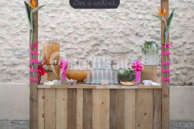 Stand en bois avec des boissons, verres et décorations tropicales, sous une pancarte ’Bar à cocktail’.