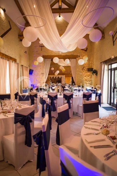Salle de réception décorée avec des tables rondes dressées, nappes blanches, chaises ornées de nœuds bleus et lanternes…
