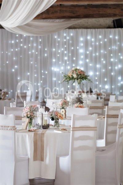 Salle de réception décorée avec des tables rondes, nappes blanches, fleurs pastel et guirlandes lumineuses en arrière-plan.