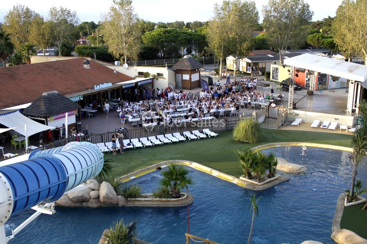 Vue d'un espace aquatique avec toboggan, piscine, chaises longues et une terrasse bondée pour un événement en plein air.