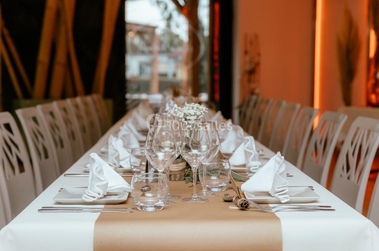 Table élégamment dressée avec nappes beiges, serviettes pliées, verres et couverts alignés dans une salle lumineuse.