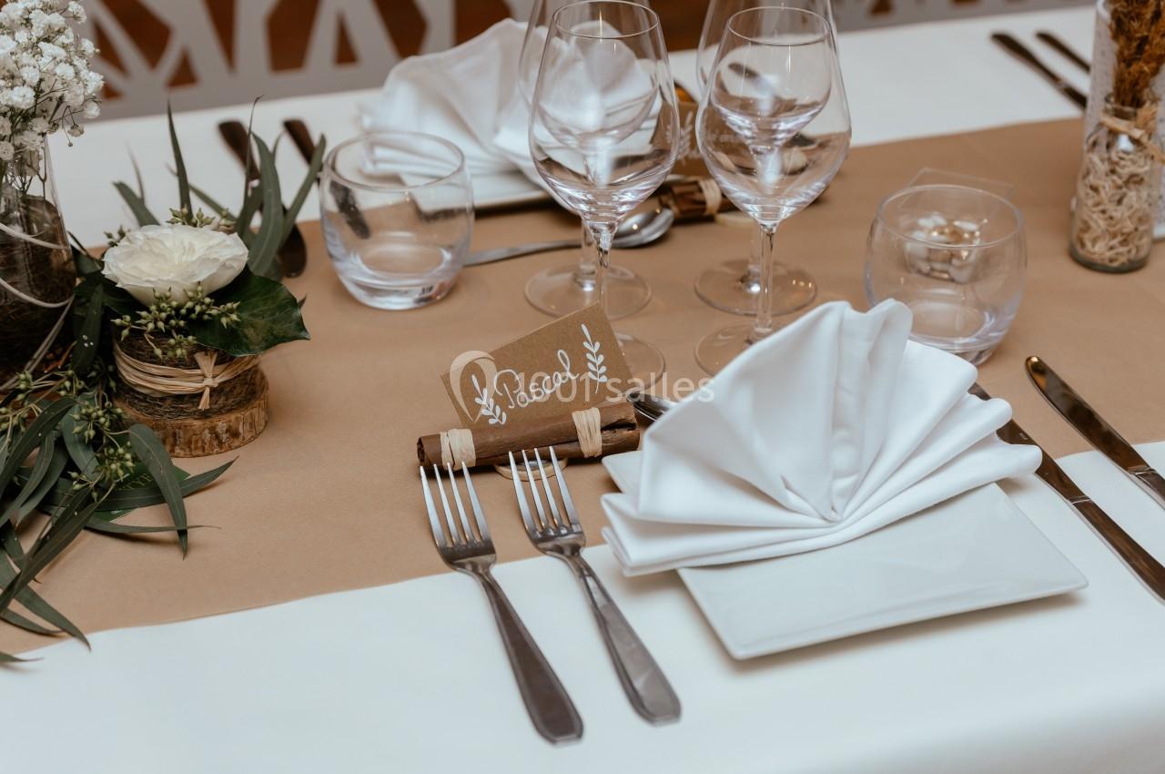 Table dressée avec nappe beige, serviette pliée, couverts, verres et marque-place ’Pascal’ décoré de feuillage.