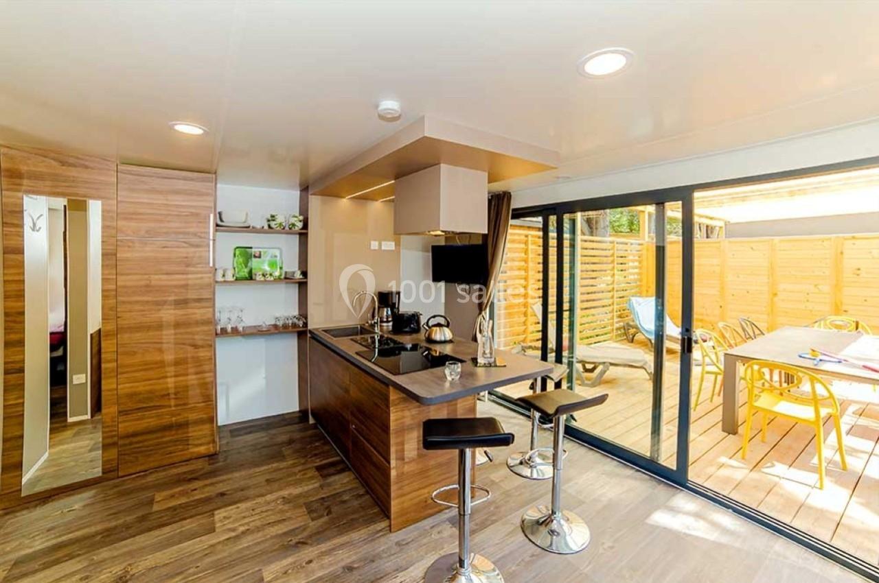 Cuisine moderne avec îlot central, tabourets hauts, et grande baie vitrée donnant sur une terrasse en bois aménagée.