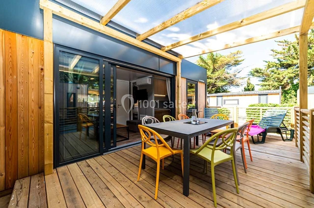 Terrasse en bois avec table, chaises colorées et pergola, donnant sur une baie vitrée ouverte vers un intérieur moderne.