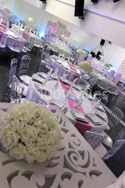 Salle de réception élégamment décorée avec des tables rondes, nappes blanches, chaises transparentes et centres de table…