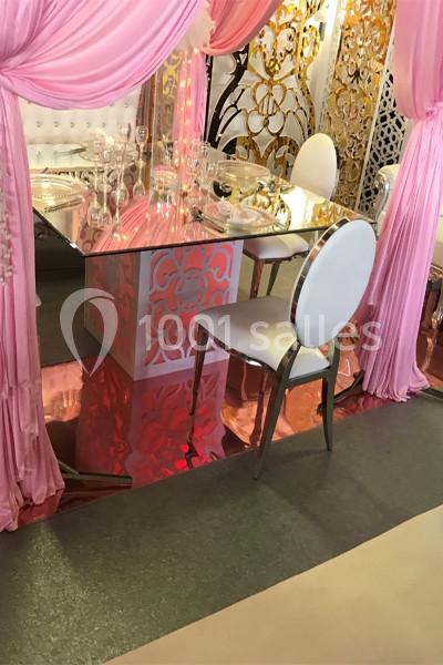 Table décorative avec nappe en verre, chaises blanches et rideaux roses, dans un cadre orné de motifs dorés.
