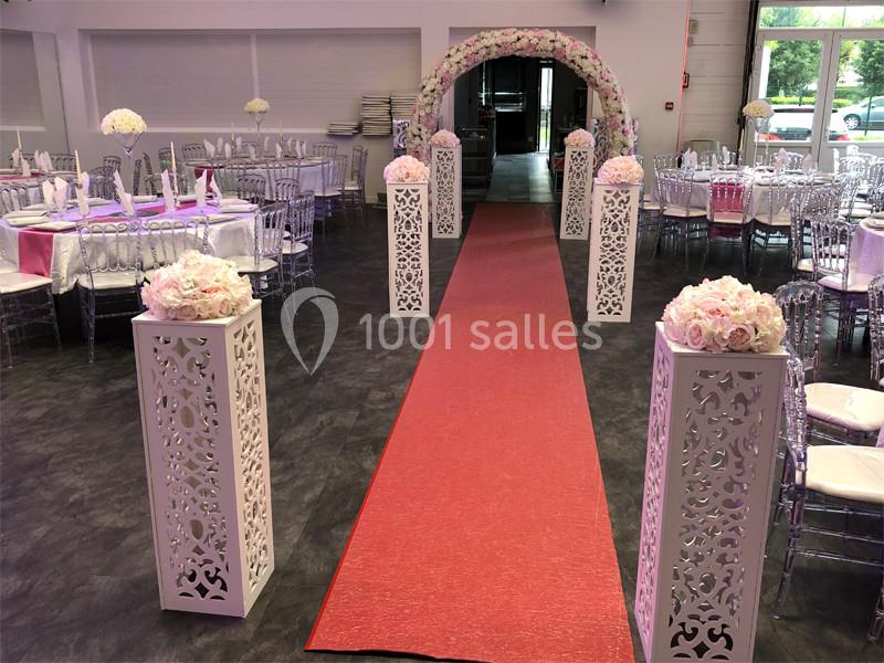 Allée centrale décorée de colonnes blanches ornées de fleurs roses, menant à une arche florale dans une salle de réception.