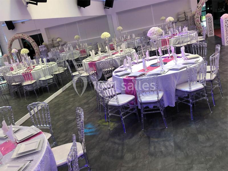 Salle de réception décorée avec des tables rondes, nappes blanches, chaises transparentes et centres de table fleuris.