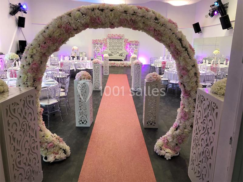 Allée centrale décorée de fleurs blanches et roses, avec arche florale et tables dressées pour un événement festif.