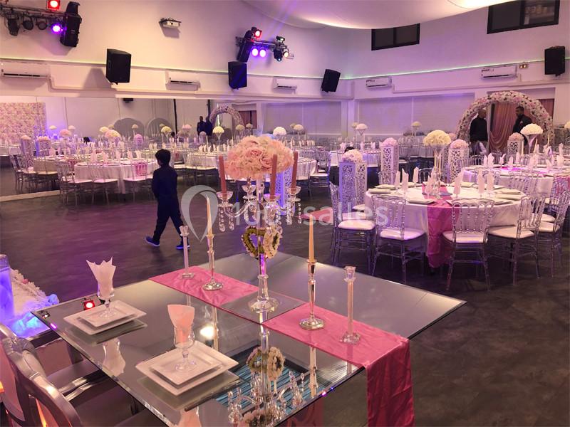 Salle de réception décorée avec des tables rondes, nappes blanches, chaises transparentes et centre de table fleuris.