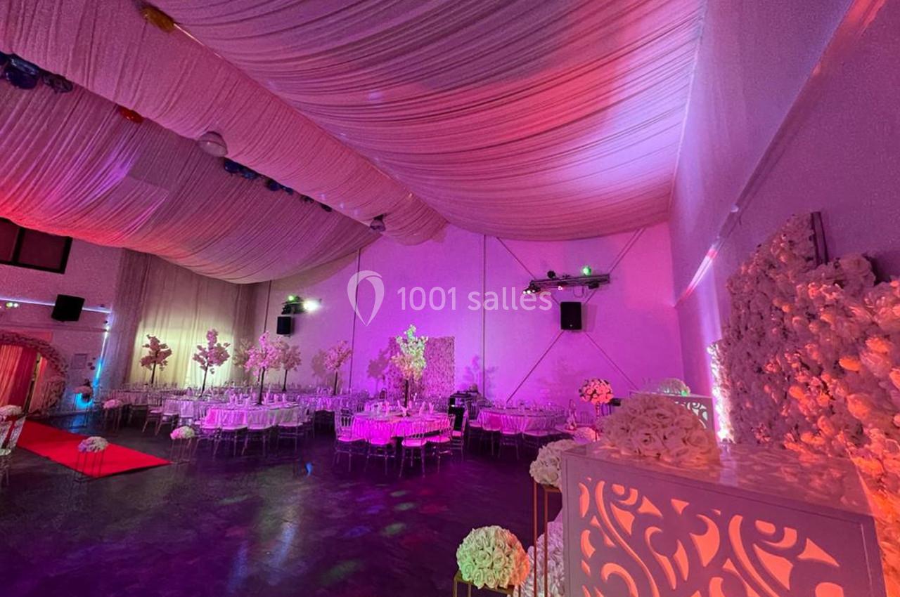 Salle de réception décorée avec des fleurs blanches, éclairages roses et violets, et tables dressées.