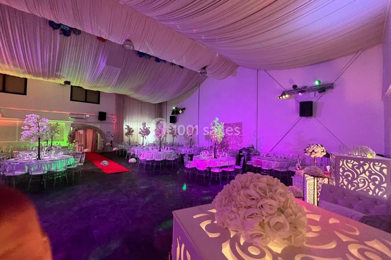 Salle de réception décorée avec des tables rondes, des chaises transparentes, des fleurs et un éclairage coloré.