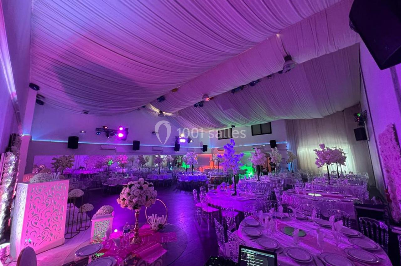 Salle de réception décorée avec des tables dressées, éclairage coloré et plafond drapé de tissu blanc.