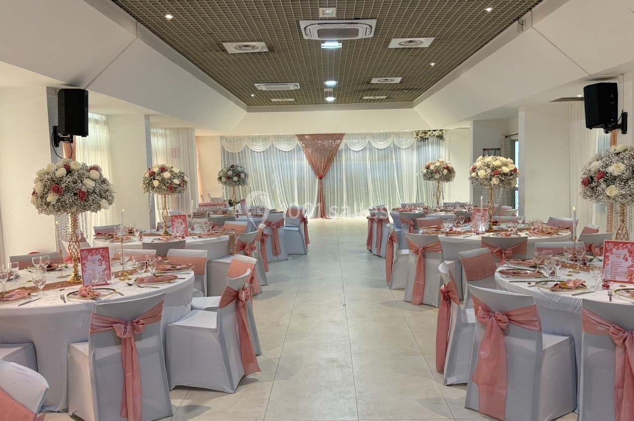 Salle de réception décorée pour un événement, avec tables rondes ornées de nappes blanches et rubans roses.