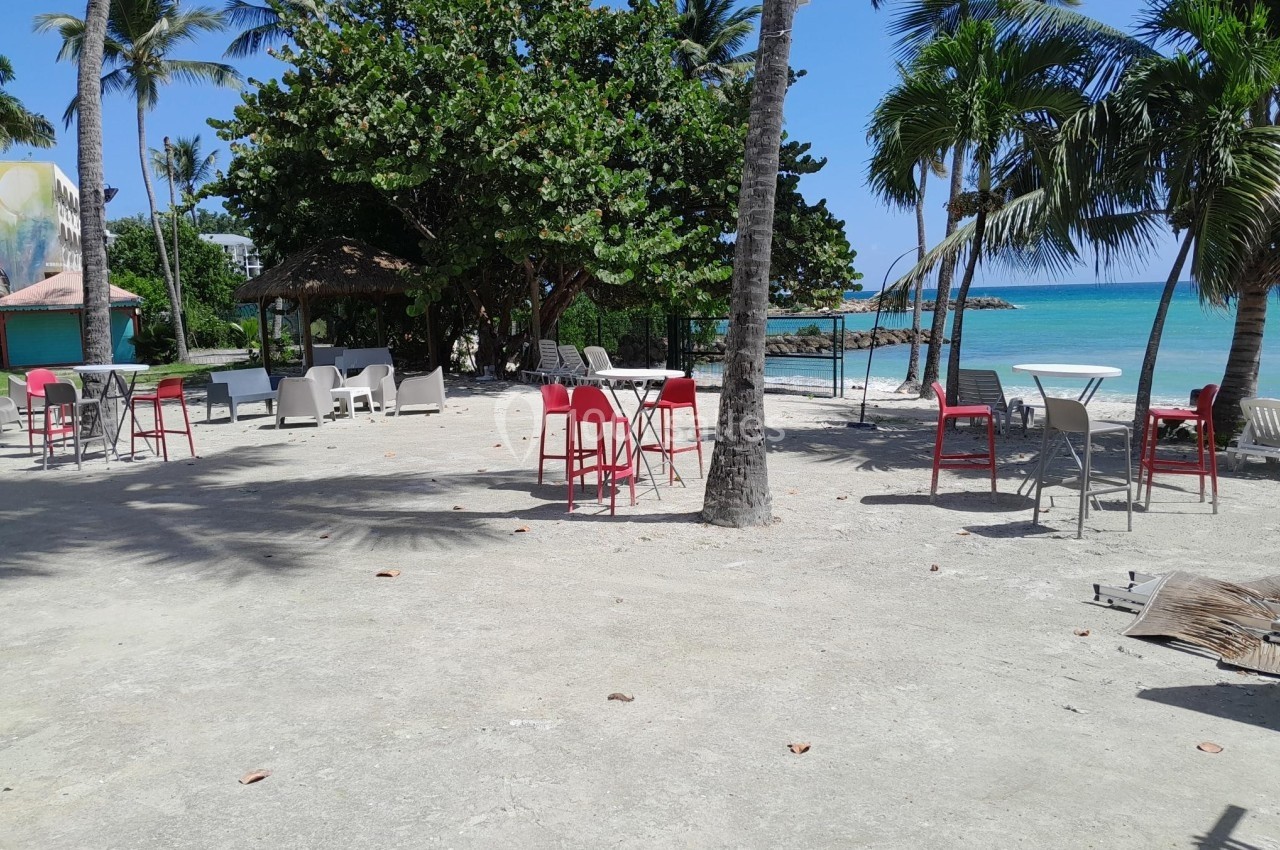 Plage avec des tables et chaises sous des palmiers, vue sur la mer turquoise et un ciel dégagé.