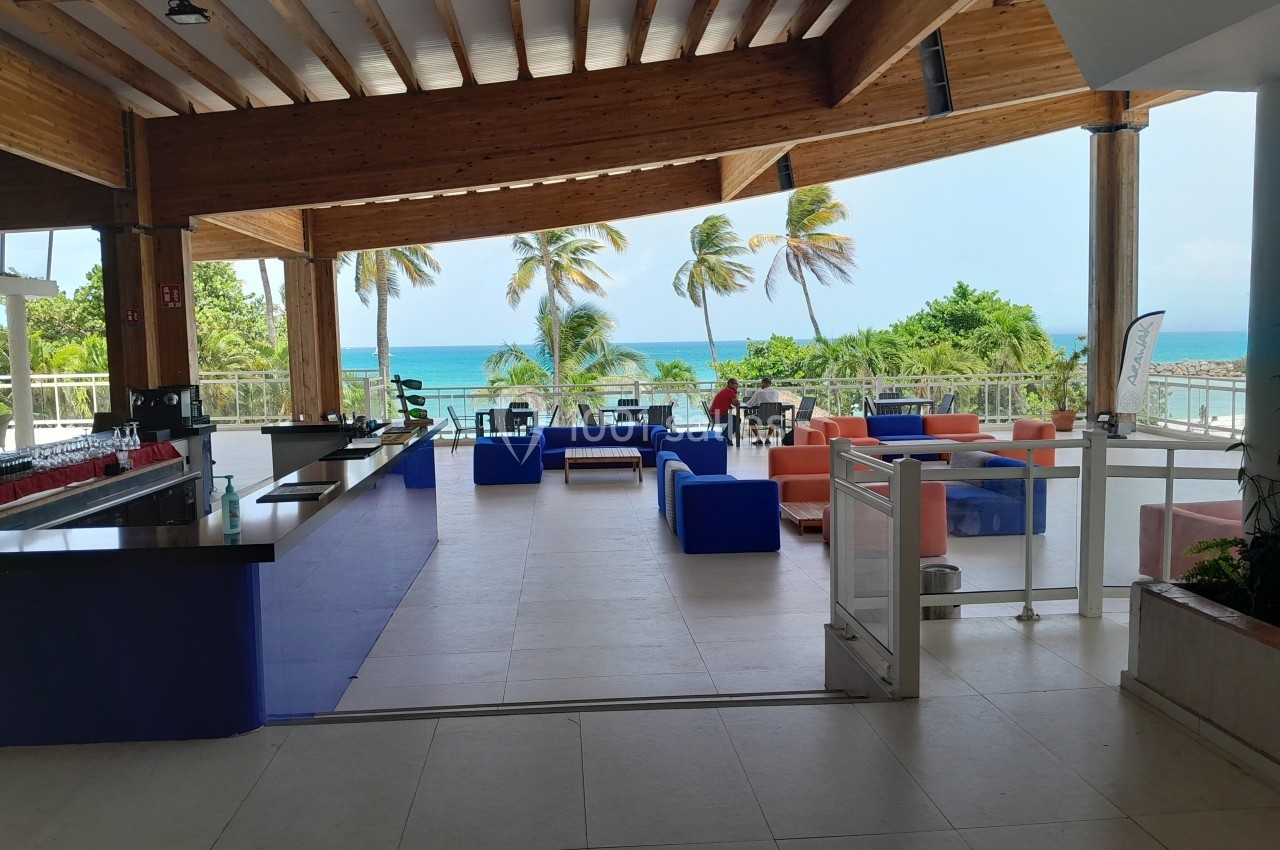 Terrasse couverte avec mobilier coloré, vue sur la mer turquoise et des palmiers sous un ciel dégagé.
