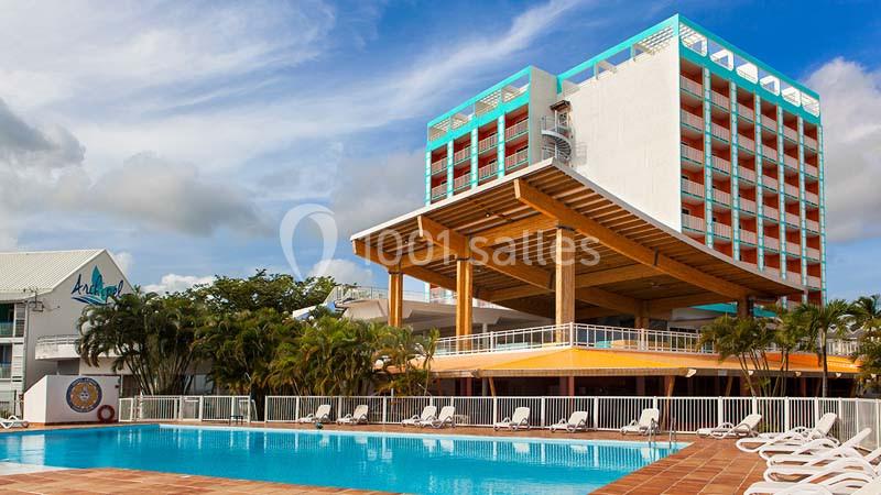 Hôtel moderne avec piscine extérieure, chaises longues et palmiers, sous un ciel partiellement nuageux.