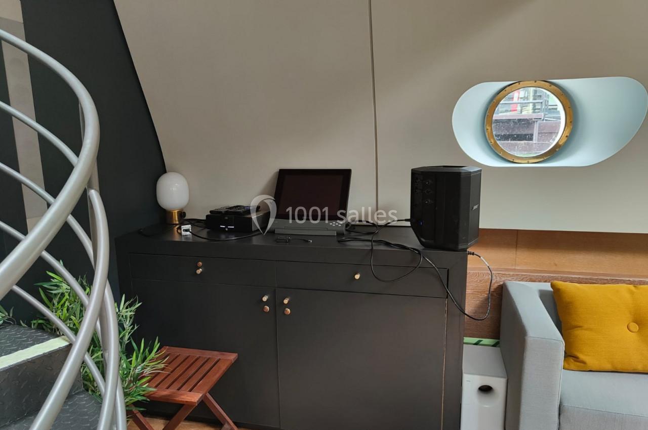 Meuble noir avec équipements électroniques, lampe, et plante, près d'un canapé gris et d'un hublot rond.