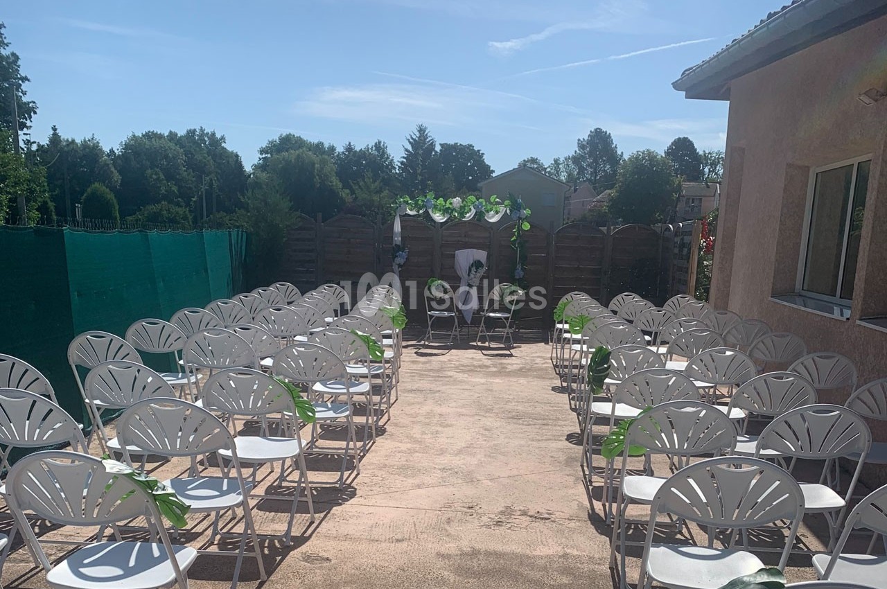 Chaises blanches alignées en extérieur devant une arche décorée de feuillage, sous un ciel ensoleillé.