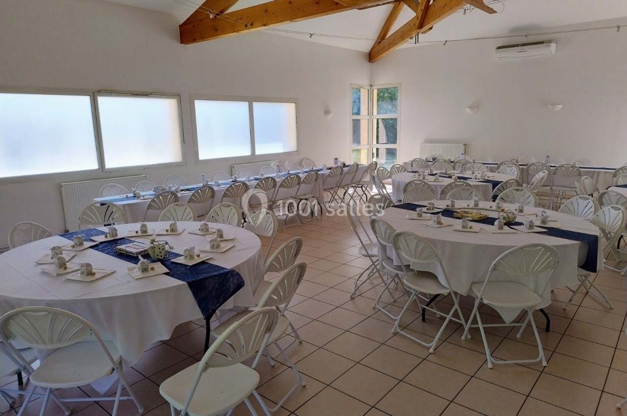Salle lumineuse aménagée avec des tables rondes et rectangulaires dressées pour un repas, chaises blanches pliantes.