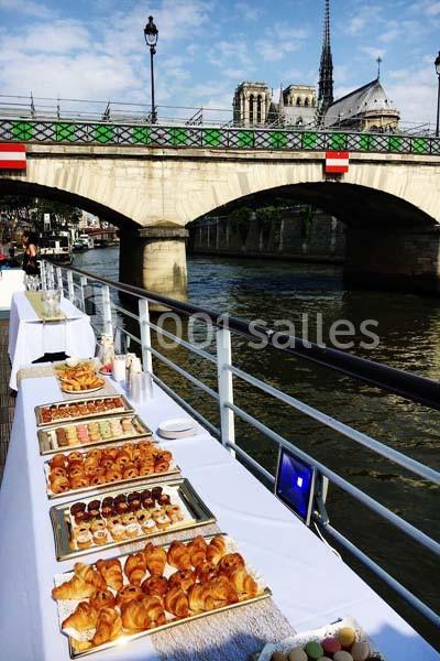 Buffet de viennoiseries et desserts sur une péniche, avec vue sur un pont et la cathédrale Notre-Dame à Paris.