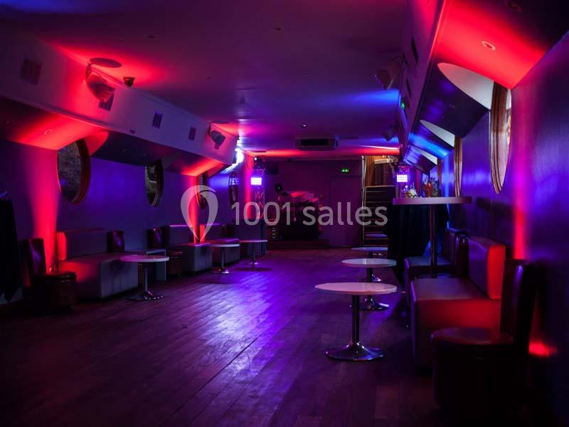 Salle de réception avec éclairage tamisé rouge et violet, tables basses et banquettes alignées le long des murs.