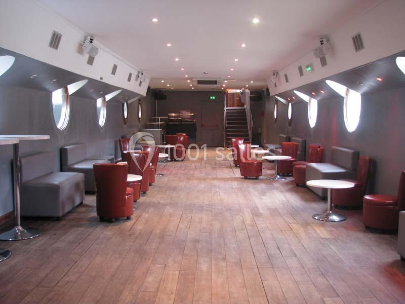 Salle intérieure avec parquet en bois, fauteuils rouges, tables blanches et hublots alignés sur les murs gris.