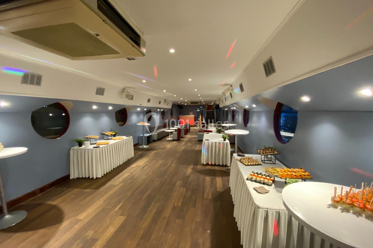 Salle aménagée avec des tables de buffet garnies, éclairage tamisé et vue sur des hublots ronds.