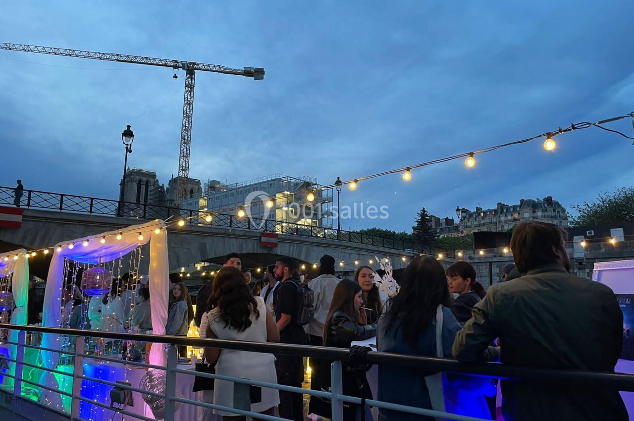 Personnes rassemblées sur une péniche illuminée au crépuscule, avec une grue et des bâtiments en arrière-plan.