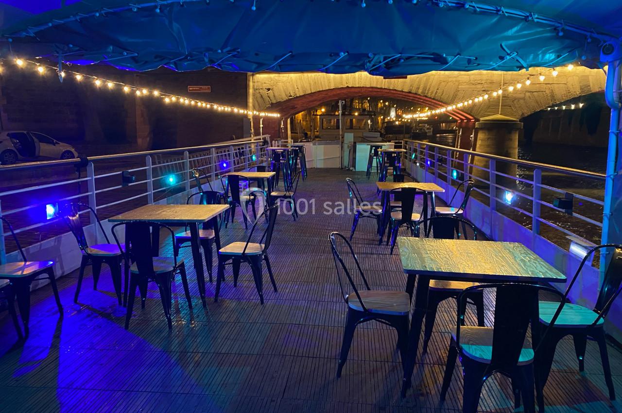Terrasse d'un bateau aménagée avec des tables et chaises sous un éclairage chaleureux, amarrée près d'un pont.