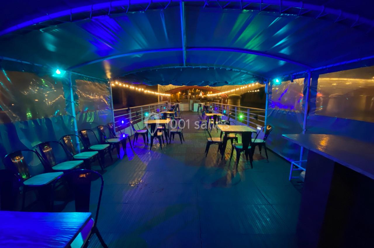 Salle extérieure d'un bateau avec tables et chaises sous un éclairage bleu et guirlandes lumineuses.